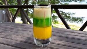 A layered Squashed Frog Shot with green, yellow, and creamy white layers, with red drops of grenadine sinking through.
